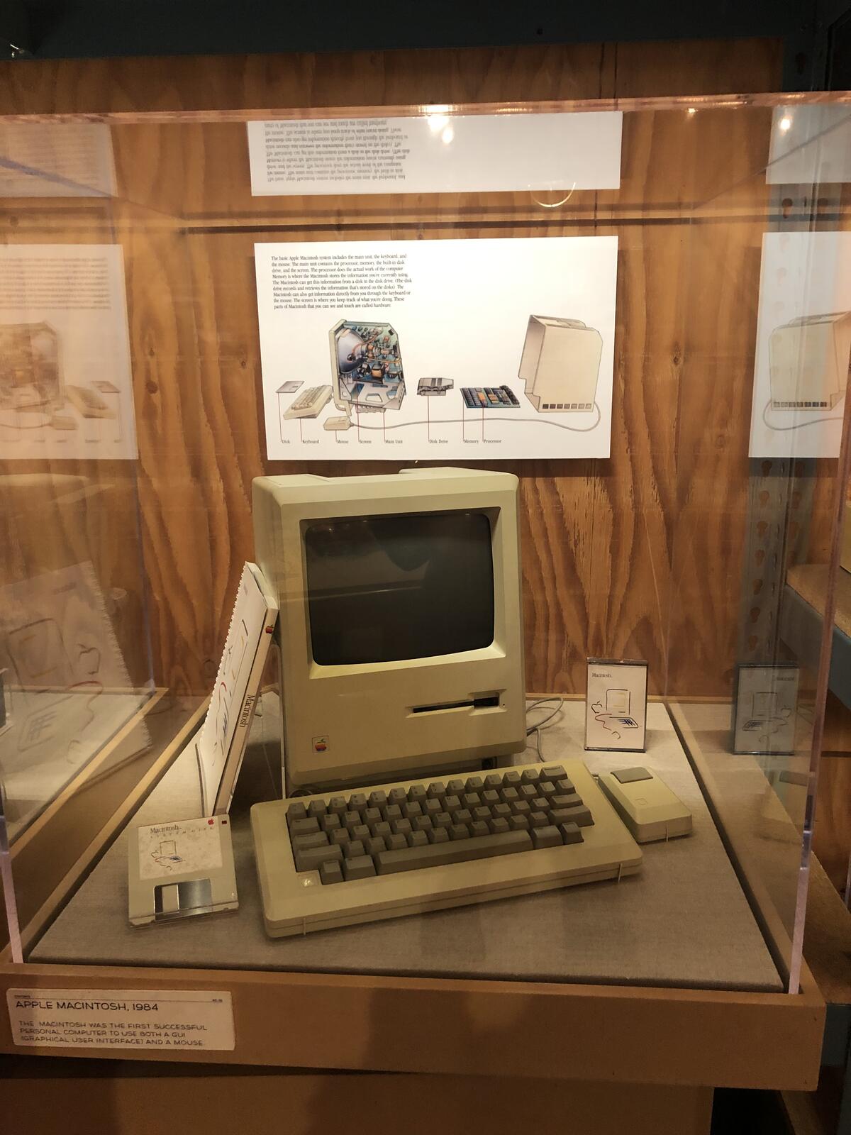 An original Macintosh in a museum display case, its manuals laid out beside it.