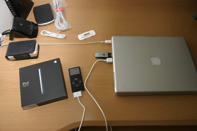 A first-generation iPod nano and its box beside a PowerBook G4 on a wooden desk, joined by a dock cable.