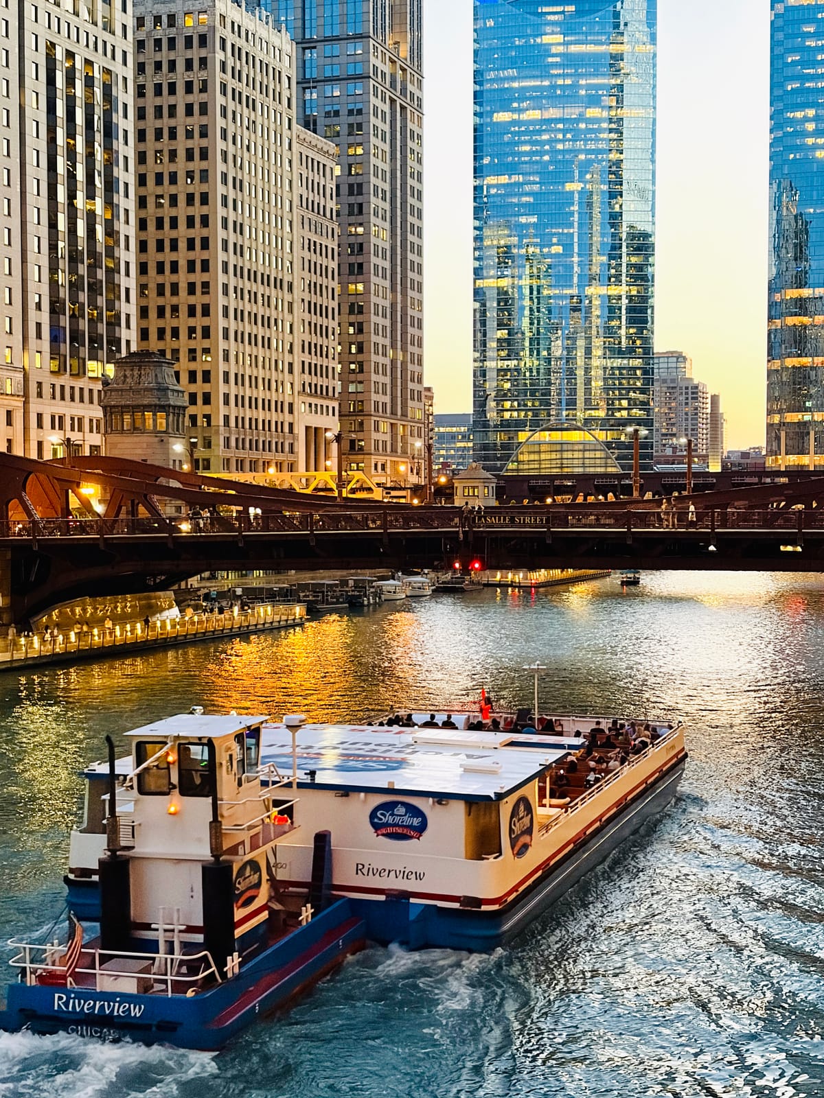 A sightseeing boat heads up the Chicago River under the LaSalle Street bridge at golden hour.