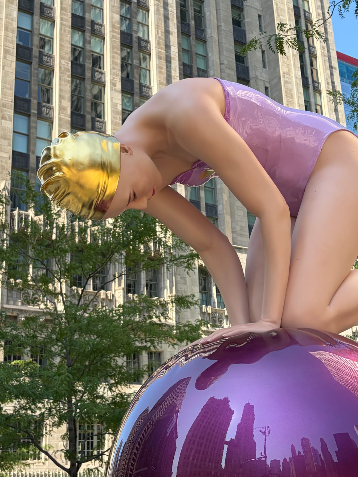 A giant swimmer in a gold cap, resting on a mirrored ball that reflects the whole skyline.