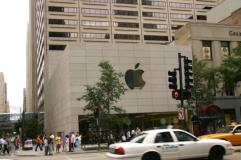 A corner store under a green apple logo, a yellow cab passing, somewhere in the US.